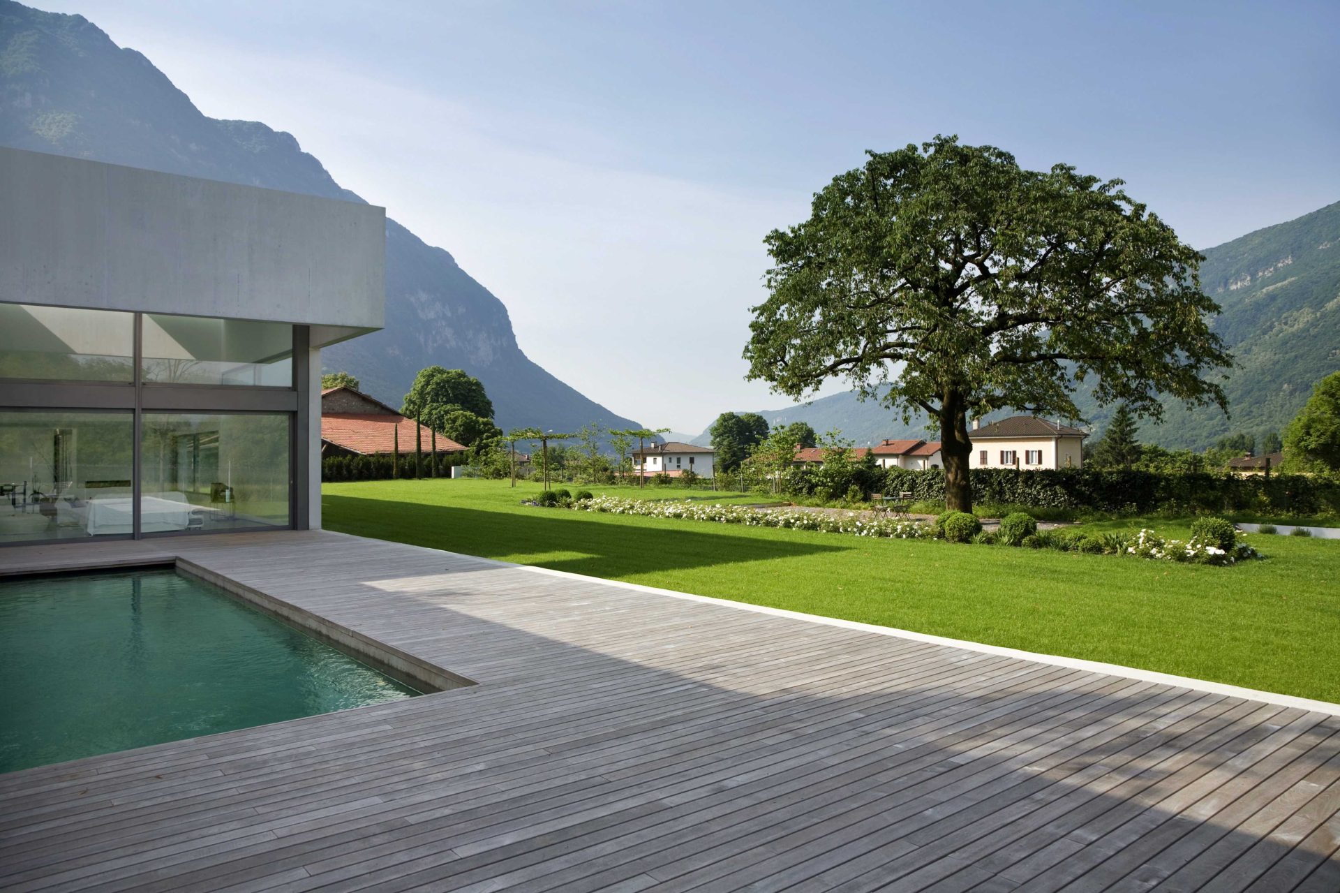 Une terrasse en bois autour d’une piscine dans un grand jardin intégré à une villa moderne aux pieds des montagnes.